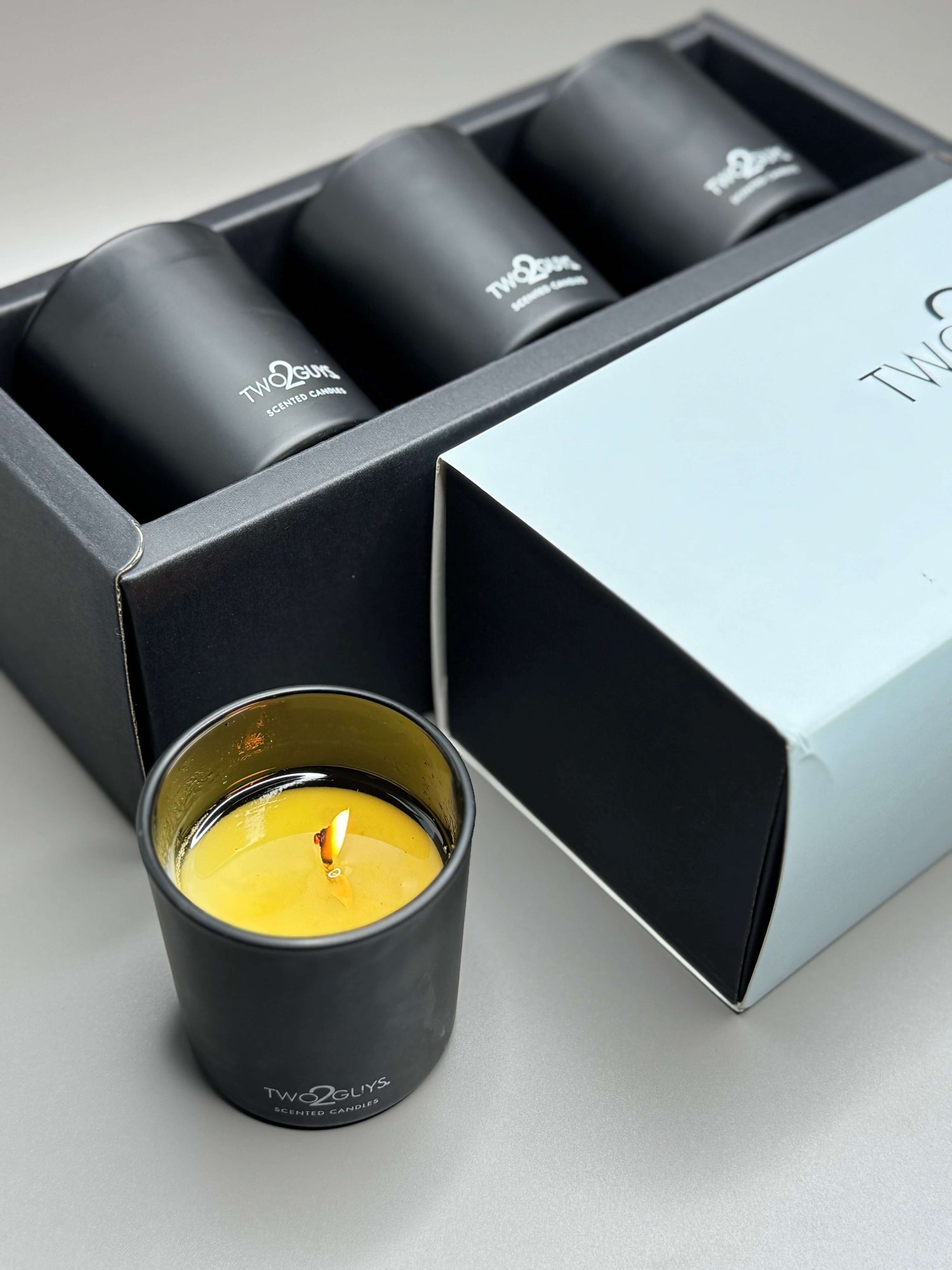 Black candle holder with a lit candle inside, placed on a gray surface with an open box in the background.