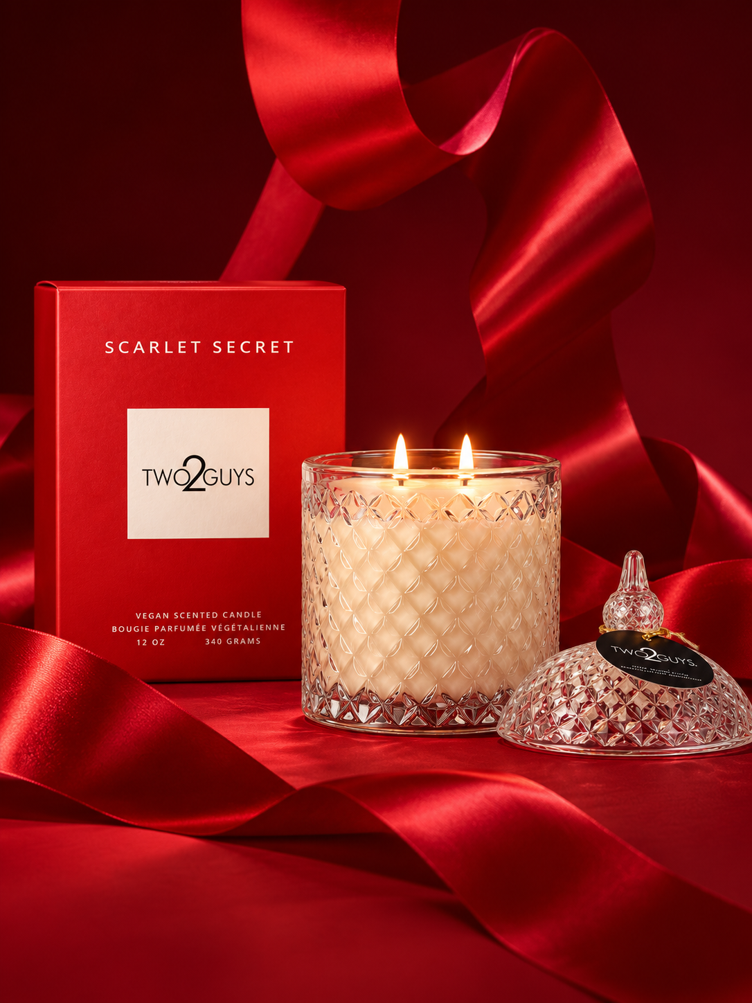 A Two Guys Scarlet Secret scented candle in a clear textured glass jar with two lit wicks, shown beside its red branded box and a matching red glass lid on a rich red satin background.