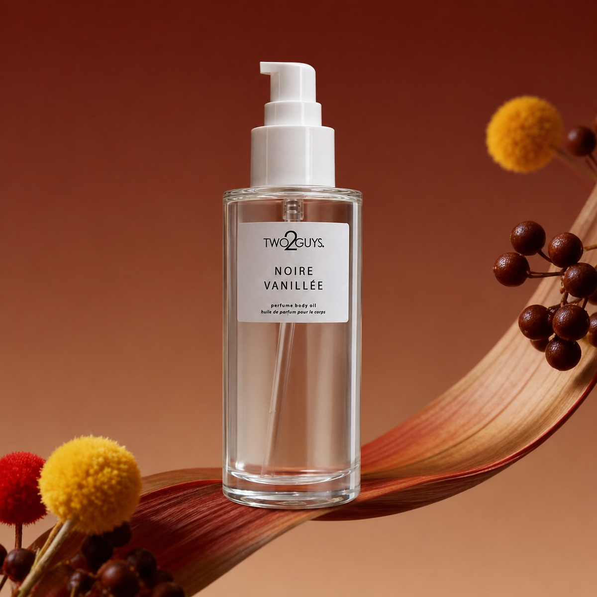 A bottle of Two Guys Noire Vanille body oil with a white pump dispenser, set against a warm brown background with decorative dried leaves and pom-pom flowers.