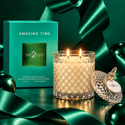 A Two Guys 'Amazing Time' scented candle in a textured glass jar with two lit flames, shown beside its teal box and matching lid on a dark green surface with teal ribbon and ornaments.