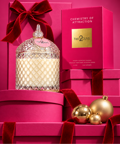 A Two Guys 'Chemistry of Attraction' scented candle in a clear patterned glass jar with a silver lid, styled on a bright pink background with matching gift boxes, red velvet ribbons, and gold holiday ornaments.