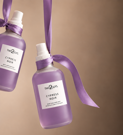 Two frosted glass spray bottles of TWEEL'S Cypress Noir hair wash with white caps and white labels, tied with purple satin ribbons, against a solid warm brown background.