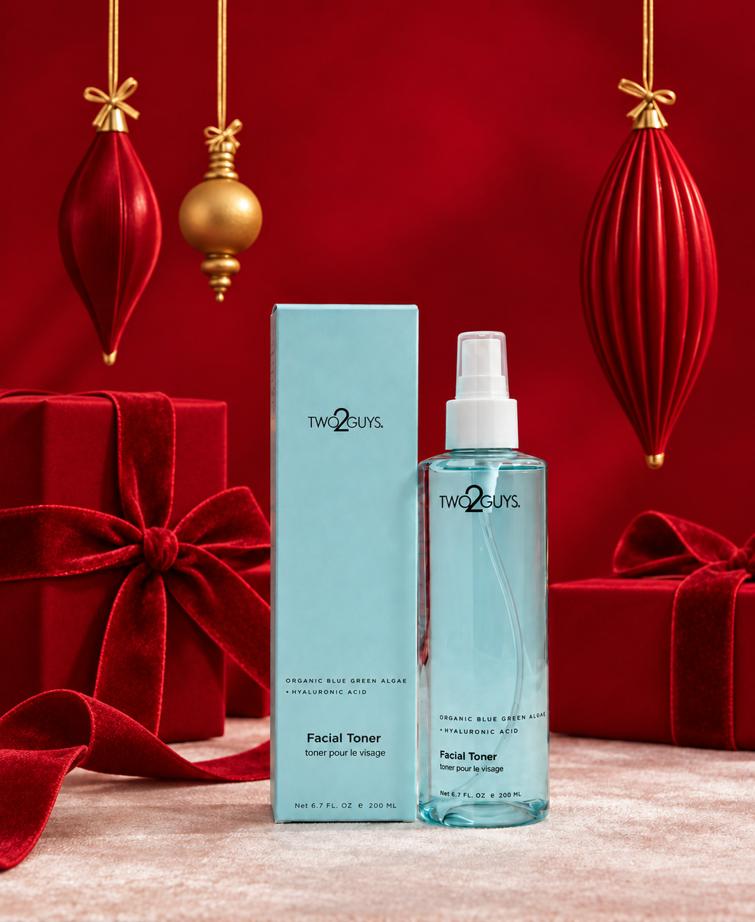 Two Guys Organic Blue Green Juice Facial Toner in a clear spray bottle with a white pump, standing next to its light blue box. The product is displayed on a beige surface with a festive red background decorated with red velvet gift boxes, ribbons, and hanging ornaments.