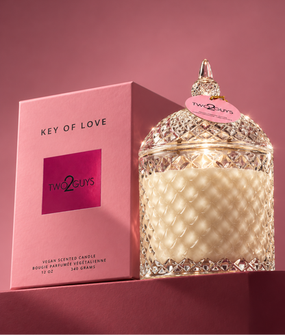 A Two2Guys 'Key of Love' scented candle in a textured glass jar with a sparkling crystal-like lid, displayed next to its pink packaging box on a matching pink background.