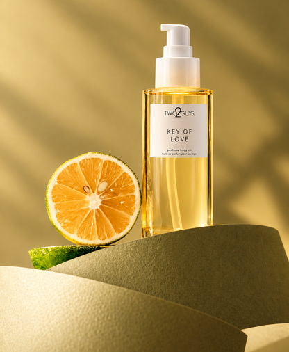 A bottle of Two 2Guys Key of Love body oil with a white pump dispenser, styled with a halved lime on a textured olive-green surface against a warm, sunlit beige background.