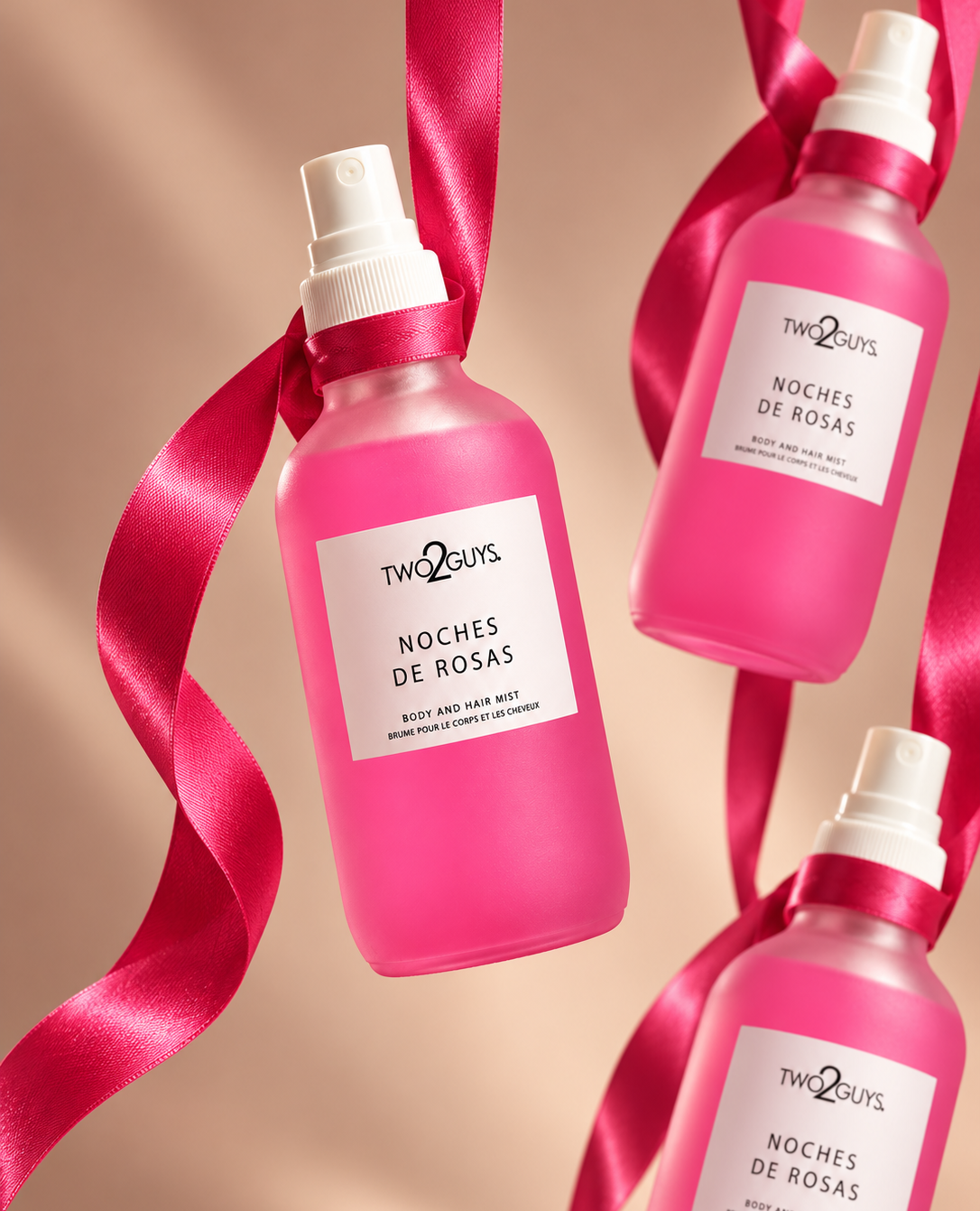 Three pink spray bottles of Two2Guys Noches de Rosas body mist, tied with bright pink satin ribbons, on a warm beige background.
