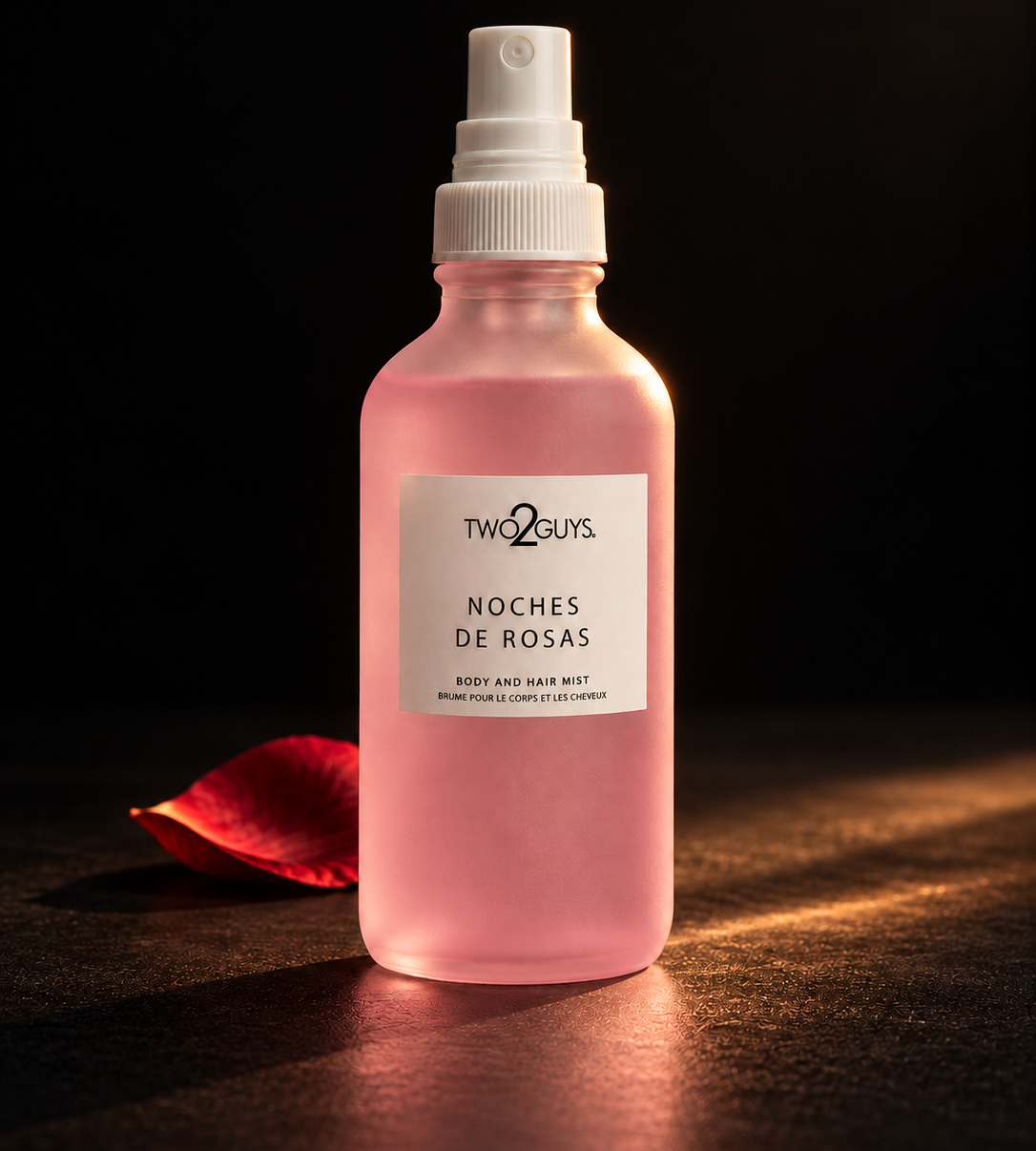 A pink spray bottle of Two Guys Noches de Rosas Body and Hair Mist, styled with a single red rose petal on a dark surface against a black background.