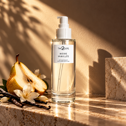 A bottle of The Scent's Noire Vanillee perfume body oil stands on a stone surface with warm sunlight and shadows. The clear glass bottle with a white pump is styled with a sliced pear, vanilla beans, and white flowers.