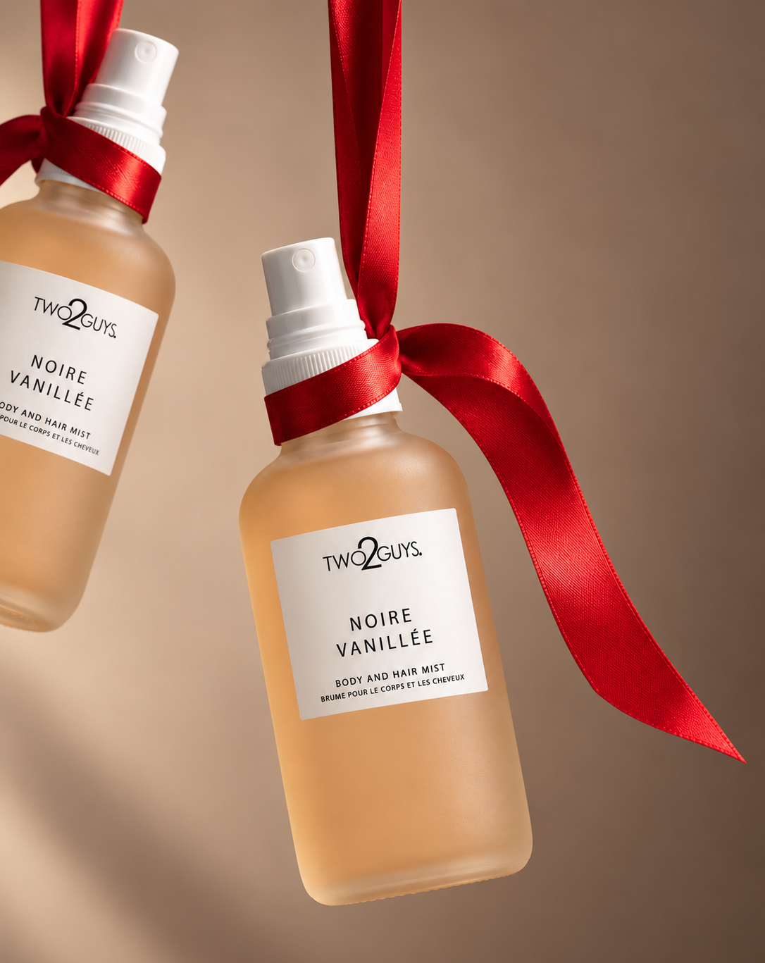 Two frosted glass spray bottles of TWO2GUYS Noire Vanille body and hair mist, tied with red ribbon and hanging against a warm beige background.