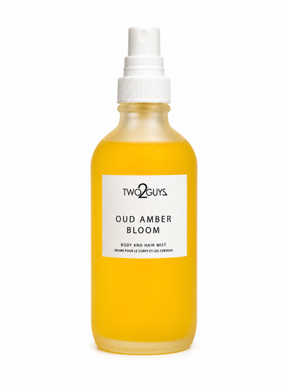 A frosted glass spray bottle filled with a golden yellow liquid, featuring a white pump dispenser and a white label with black text that reads 'TWO2GUYS OUD AMBER BLOOM'.