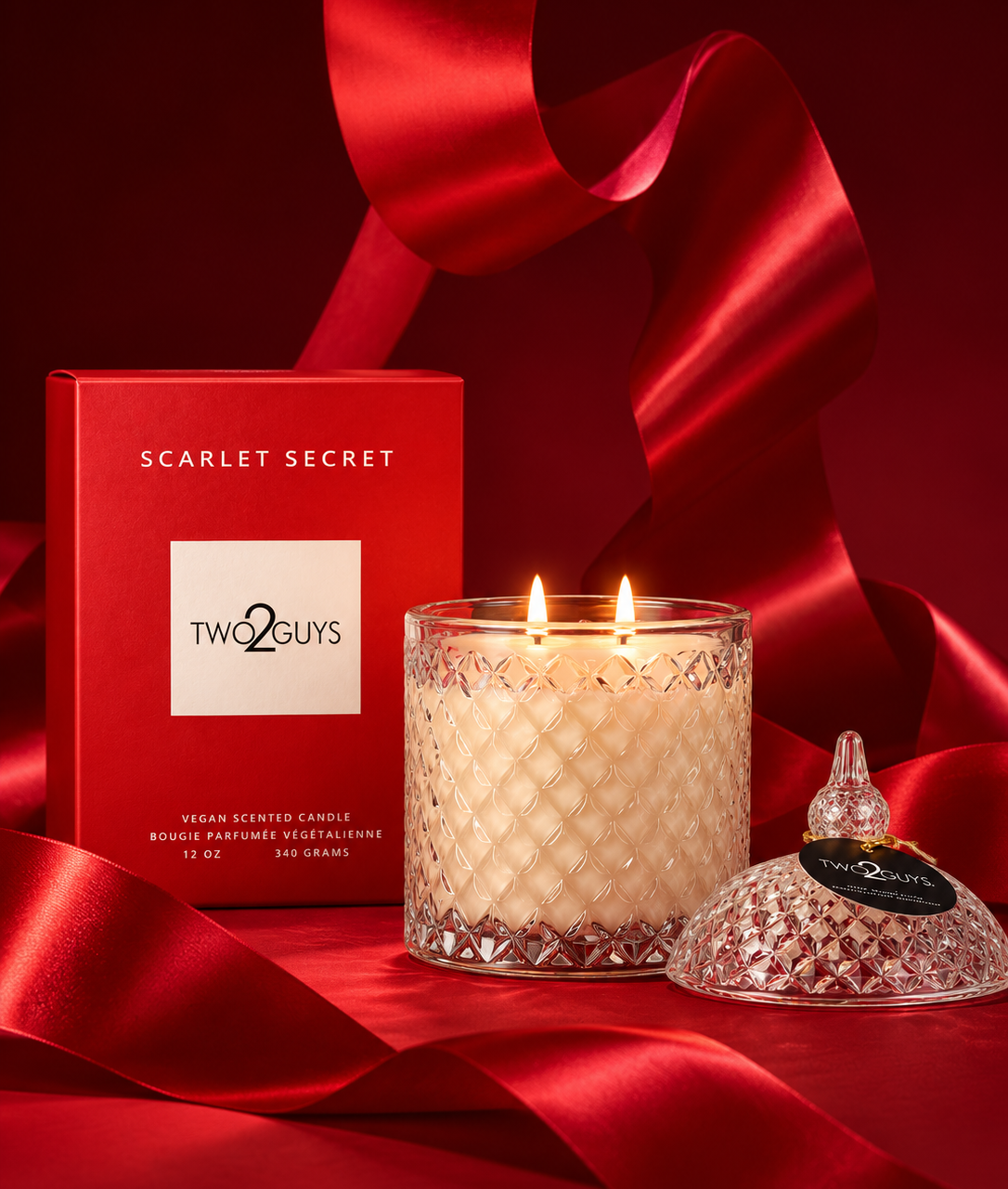 A Two Guys Scarlet Secret scented candle in a clear textured glass jar with two lit wicks, shown beside its red branded box and a matching red glass lid on a rich red satin background.
