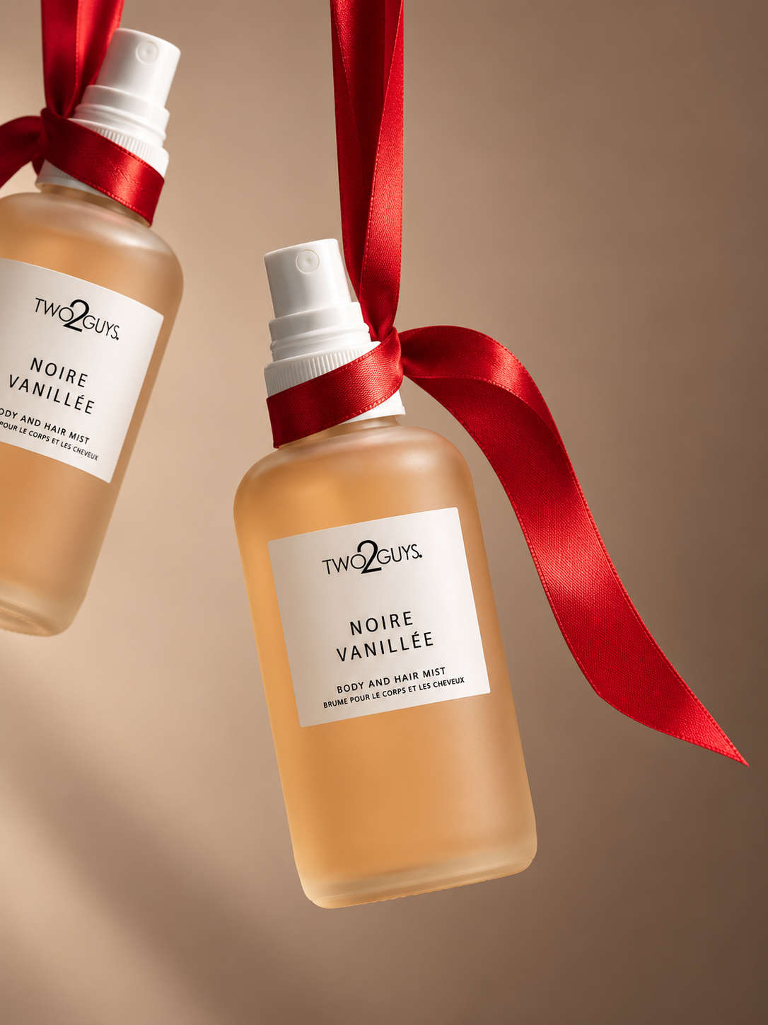 Two frosted glass bottles of Two Guys Noire Vanillee body and hair mist, decorated with red ribbons and hanging against a warm beige background.